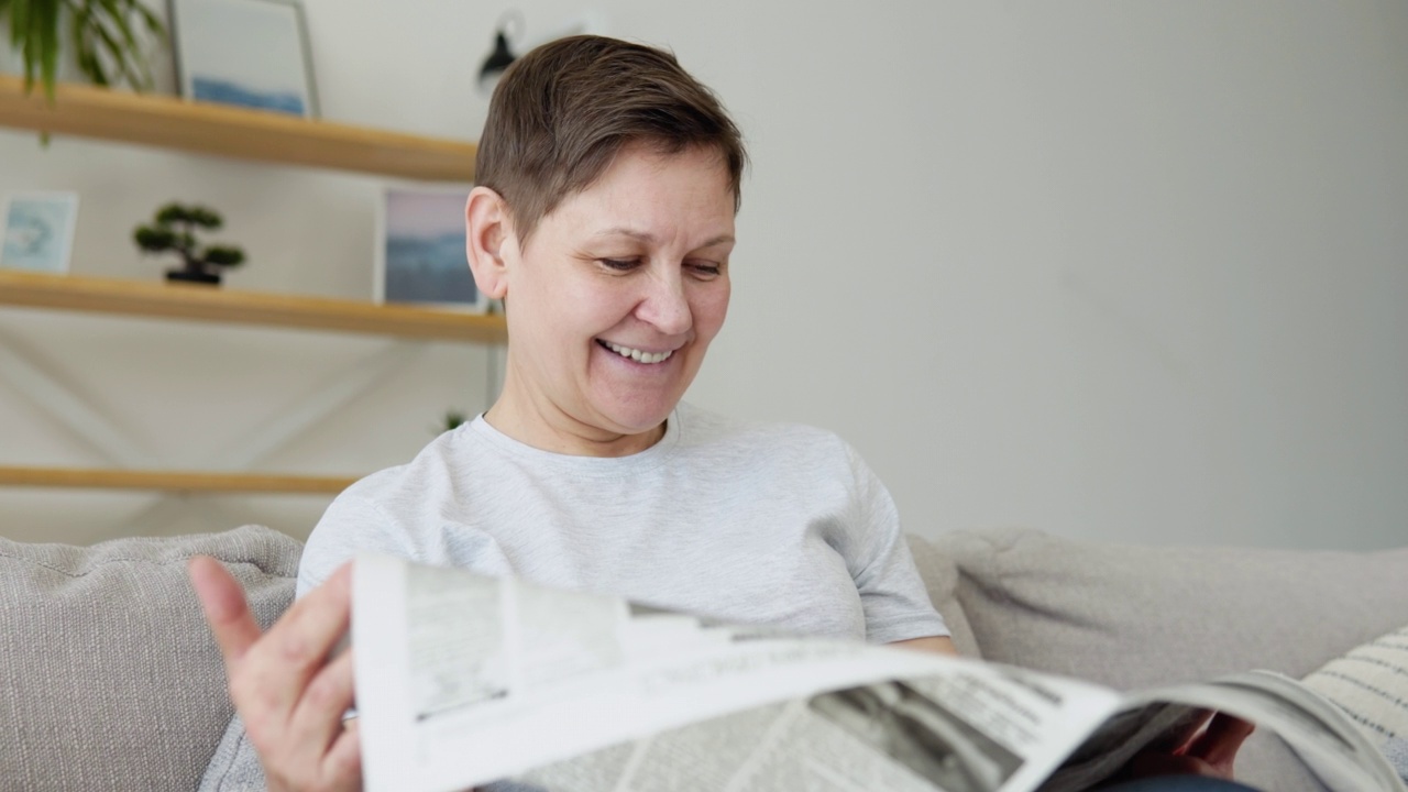 一个美丽成熟的女人坐在家里的沙发上看报纸的特写镜头。高级妇女读报纸的肖像视频下载