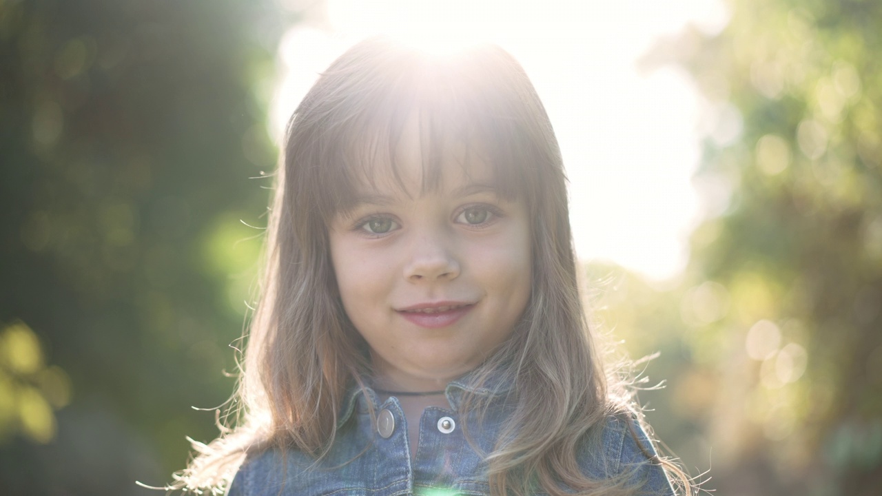 迷人快乐的白人小女孩在户外阳光摆姿势的头像。可爱的孩子微笑着看着相机站在阳光与玩具在春夏公园。缓慢的运动。视频下载