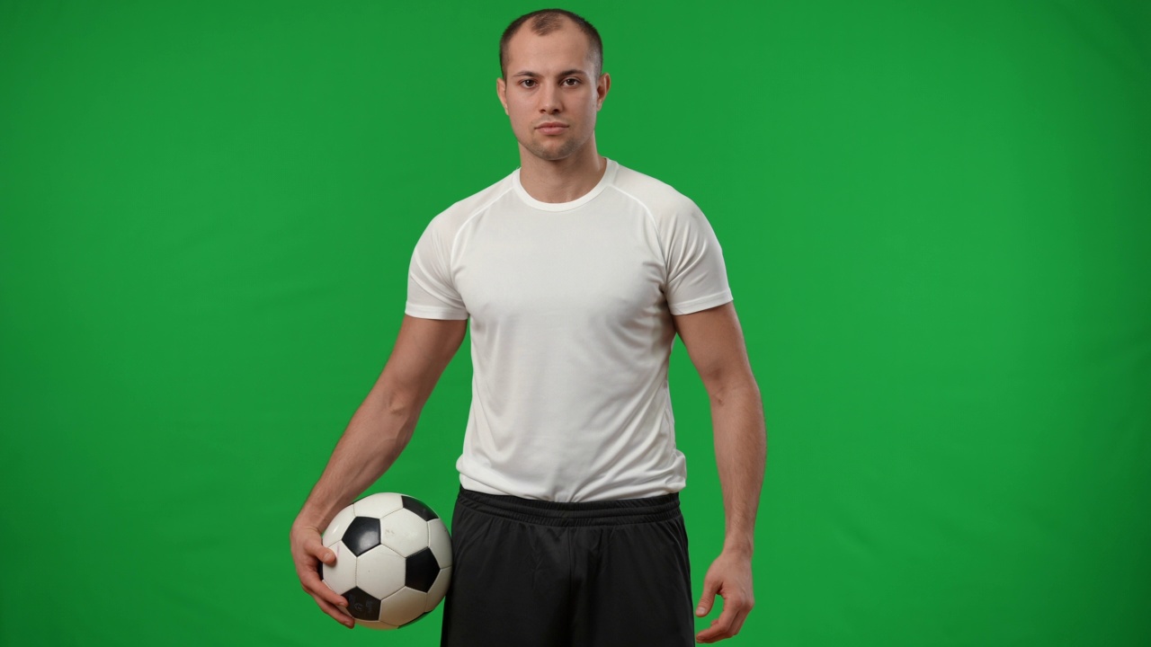 一个自信的白人年轻人在绿色屏幕上看着镜头，用慢动作捕捉足球。激励激励的运动员在chromakey背景摆姿势的肖像。视频素材