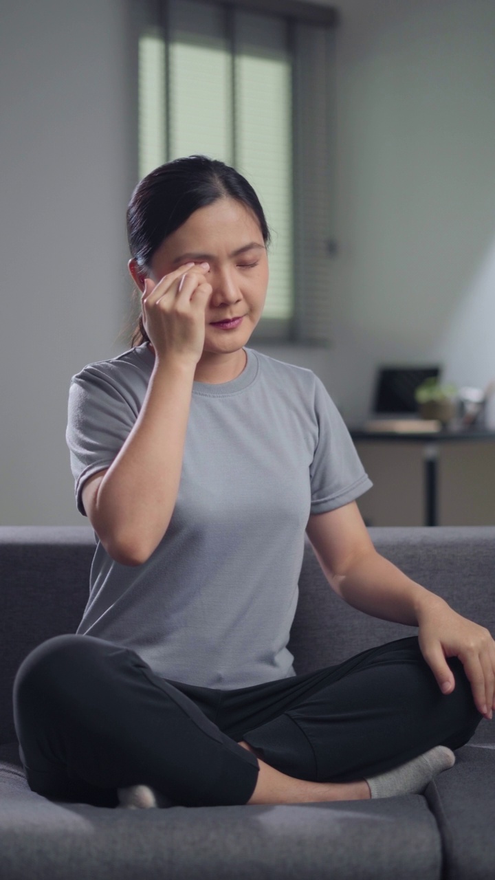 一名亚洲妇女坐在家里的沙发上，眼睛疼得难受。视频素材