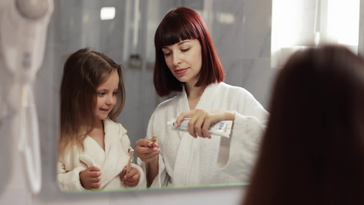 微笑的妈妈和她可爱的小女儿穿着白色浴袍在牙刷上挤牙膏视频素材