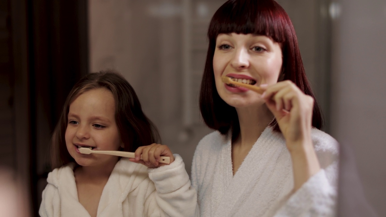 妈妈和穿着白色浴袍的可爱女儿站在镜子旁边刷牙。视频素材