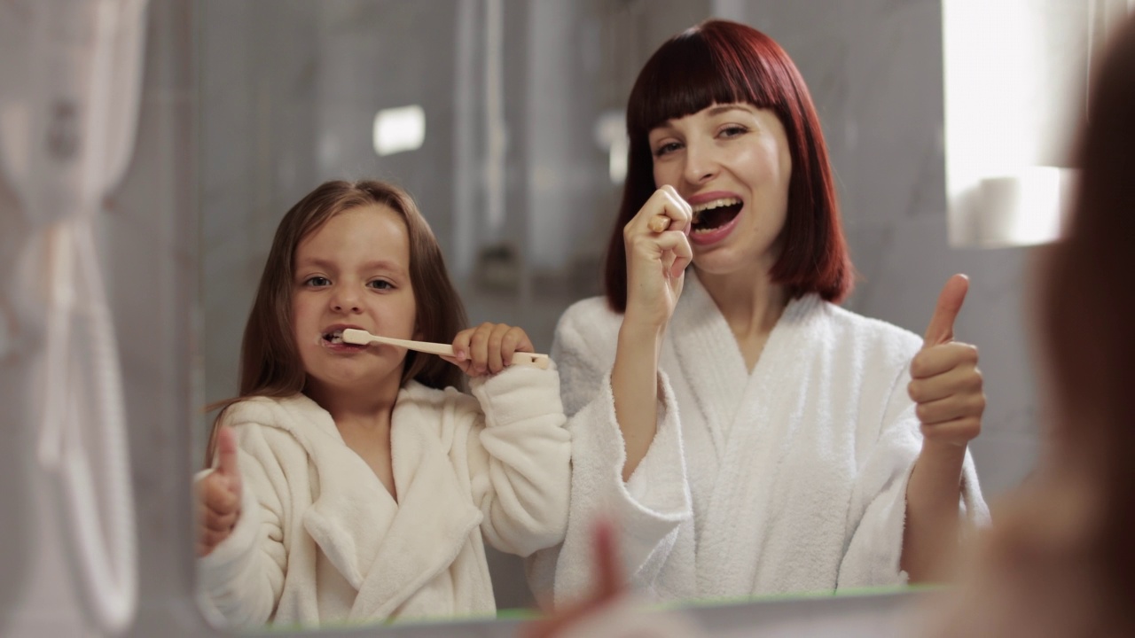 美丽的年轻妈妈和她可爱的女儿在白色浴袍刷牙的特写视频素材