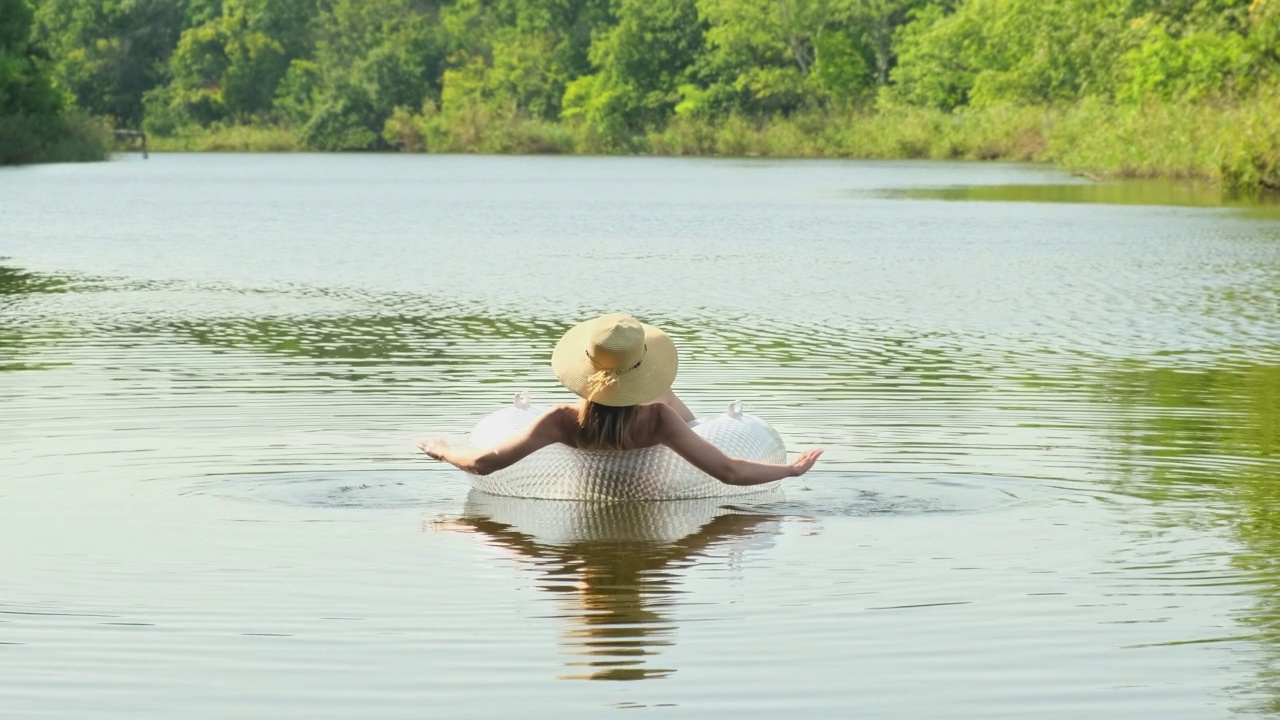 身穿泳装、头戴草帽的女子带着一个充气圆圈漂浮在湖河塘中。女孩躺在碧水的游泳池里的游泳圈上。旅游的概念，假期，周末，节假日，在大自然中放松，休息，放松视频下载
