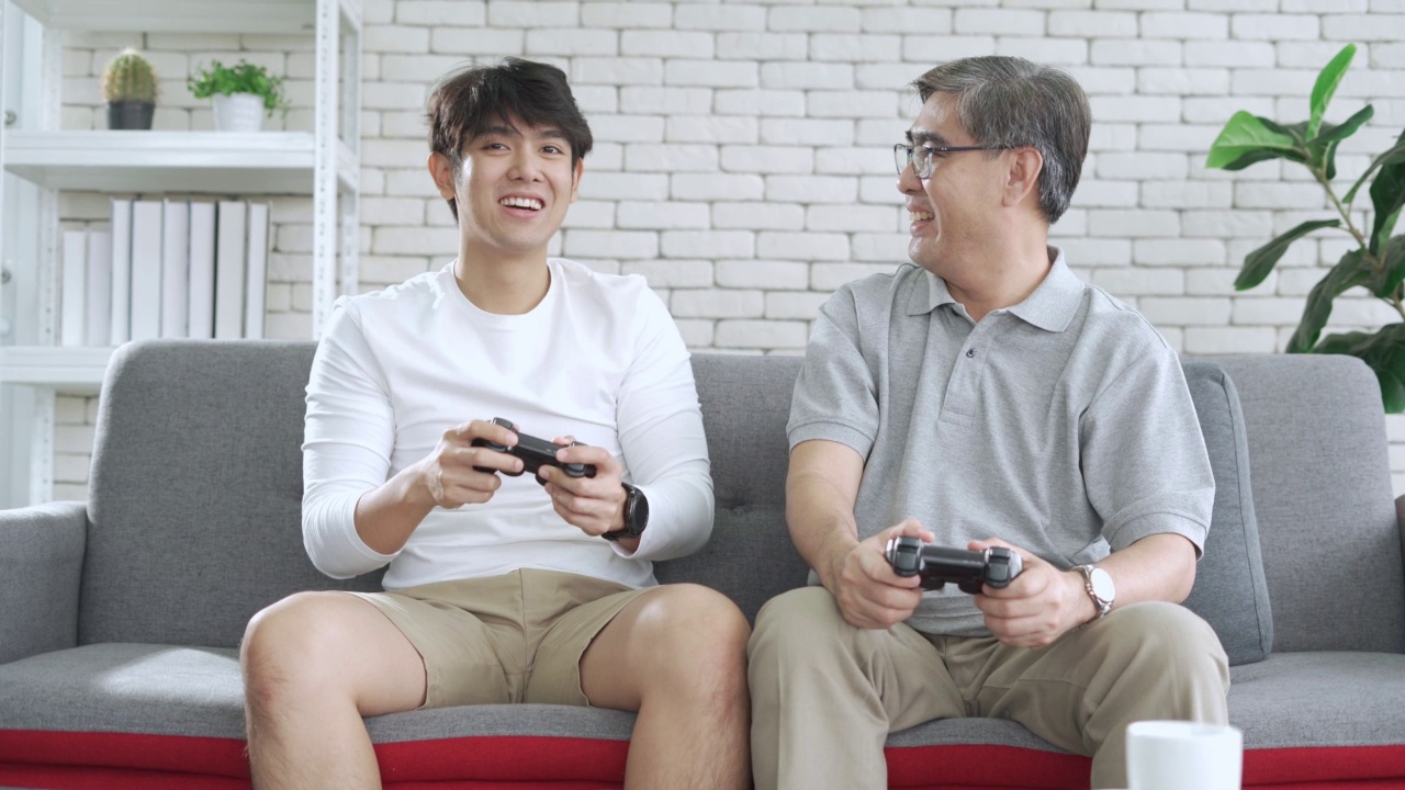 亚洲资深父亲和他的成年儿子喜欢在家里的客厅一起玩电子游戏，享受电子游戏，父母，家庭视频素材