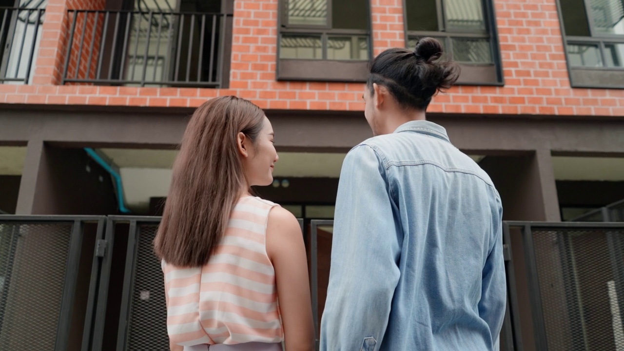 年轻的亚洲夫妇抱着对方站在后面的观点，看在新家前面。考虑买房后，一对迷人的夫妇在新房子前拥抱。视频下载