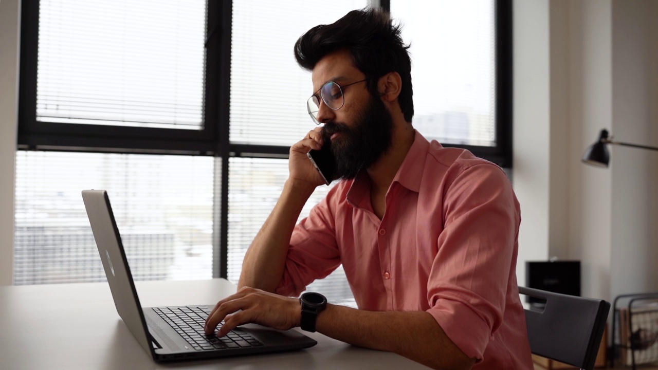 开朗的印度商人戴着眼镜在笔记本电脑前工作，坐在窗口背景的桌子上用手机聊天。视频素材