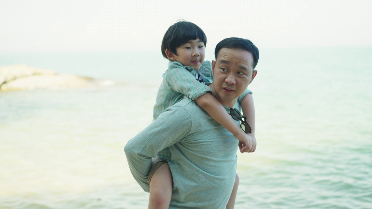 亚洲父亲背着他的小儿子，愉快地沿着海滩散步，并指出一些有趣的事情。与家人共度美好的假期时光。视频下载