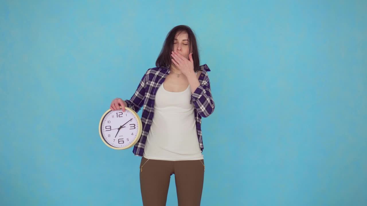 画像中昏昏欲睡的年轻女子在早晨打哈欠，手里拿着一个大钟，看着相机孤立在蓝色背景上视频下载