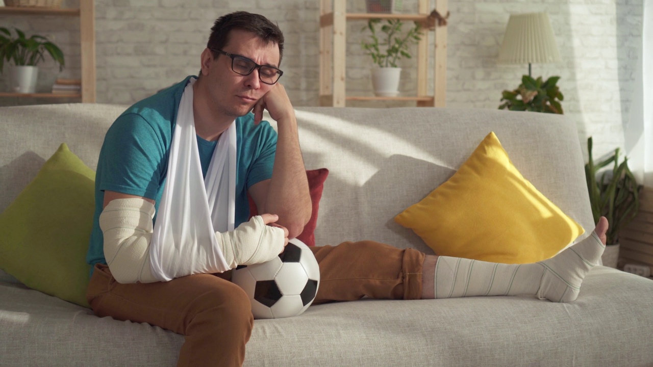 肖像伤心受伤的运动员和一个足球坐在沙发上的现代公寓视频素材