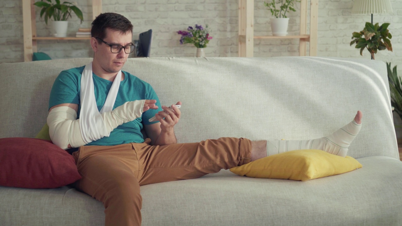 一个断了胳膊和腿的年轻男子的肖像坐在一个现代公寓的沙发上，使用智能手机视频素材