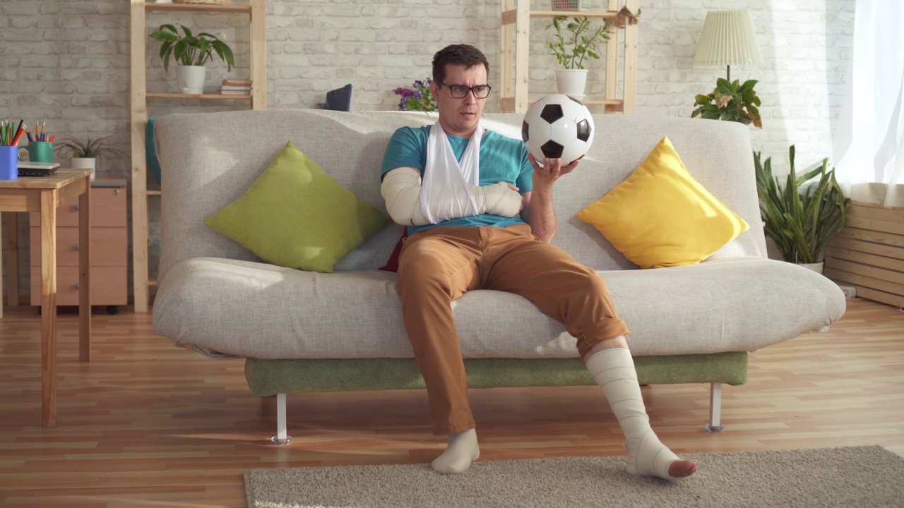 伤心受伤的运动员拿着足球坐在现代公寓的沙发上视频素材