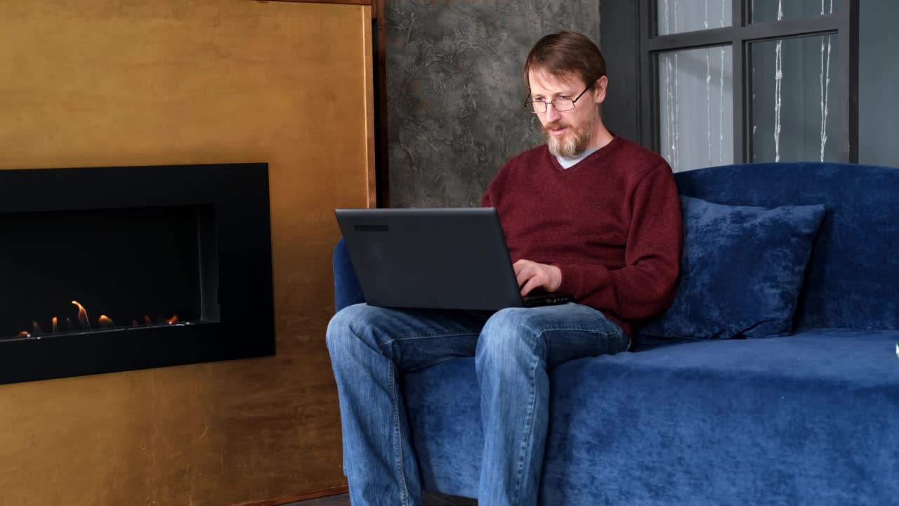 快乐的大胡子在办公室工作在家远程工作。一个五十岁的男人在壁炉旁使用笔记本电脑。在线教育与工作视频下载