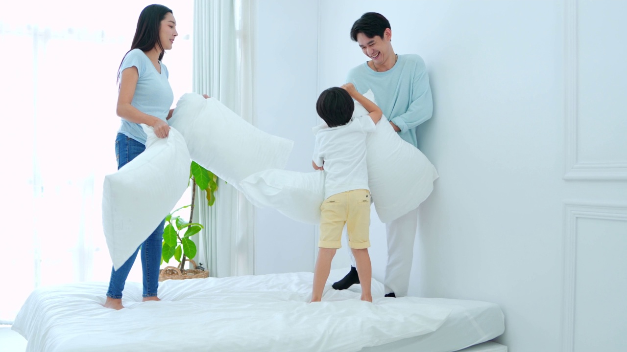 快乐的家庭时间，年轻的亚洲父母正在床上玩枕头与儿子在卧室快乐，笑着微笑。视频素材