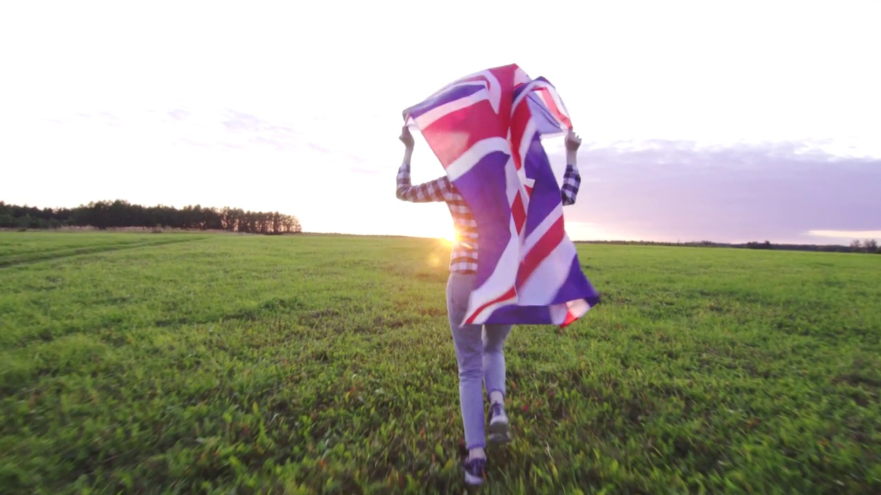 年轻积极的女人在日落时穿着衬衫，拿着英国国旗跑过田野视频下载