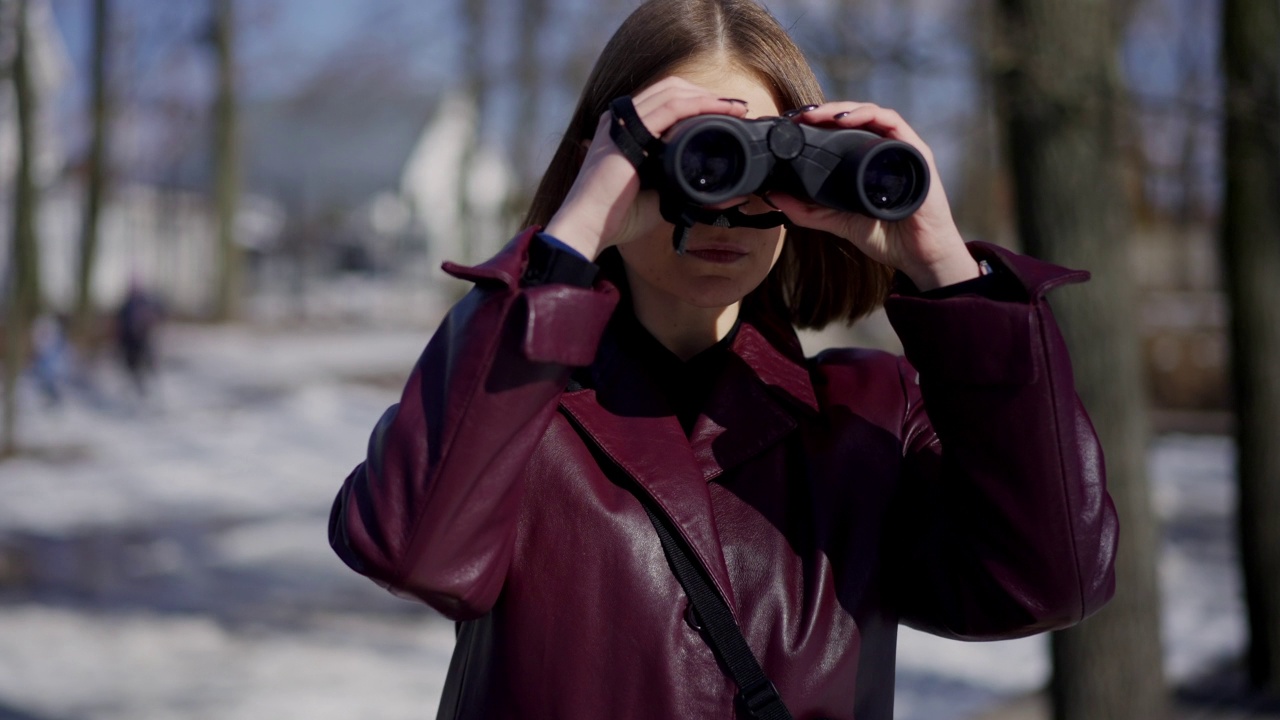 肖像严肃的女人与双筒望远镜站在冬季公园望向别处。直播镜头放大到自信苗条的高加索私家侦探调查案件侦查户外晴天。视频下载
