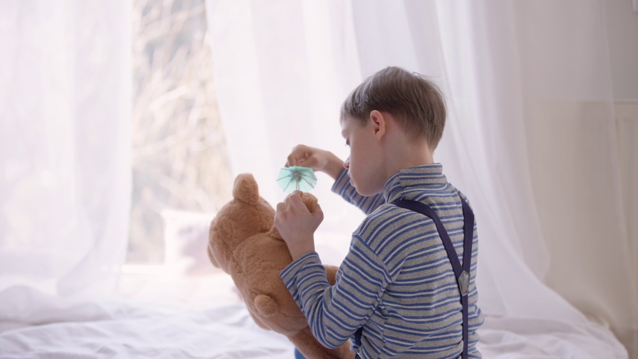 从肩膀上方拍摄一个自闭症男孩在家里的卧室里玩鸡尾酒伞和泰迪熊。愉快放松的精神障碍白人儿童坐在床上享受休闲。视频素材