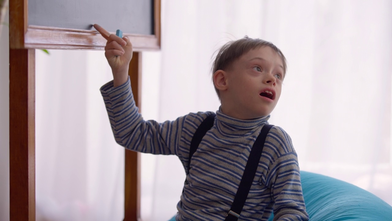 古玩自闭症男孩坐在教室里的书包椅上，指着黑板听。出生异常的白人孩子在学校室内学习的画像。教育与自闭症概念。视频下载