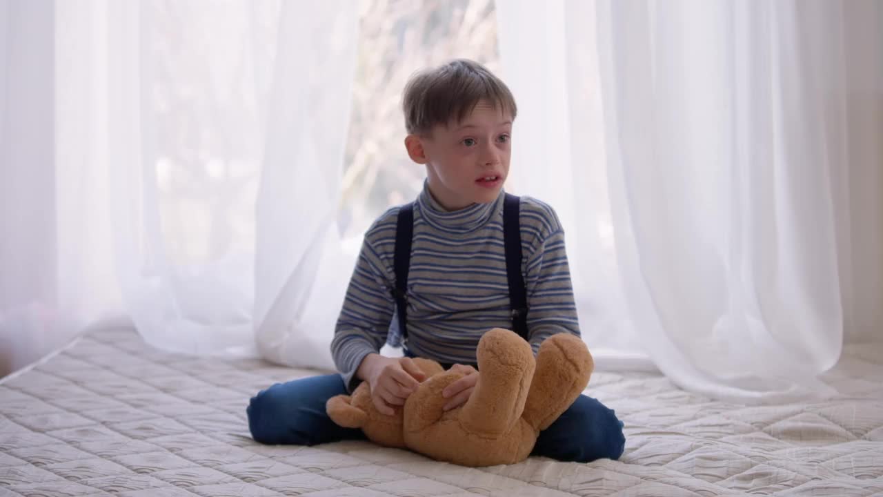 自闭症男孩带着泰迪熊坐在家里的床上，伸出舌头玩耍。放松古玩肖像与出生异常白人儿童在室内卧室休息。视频素材