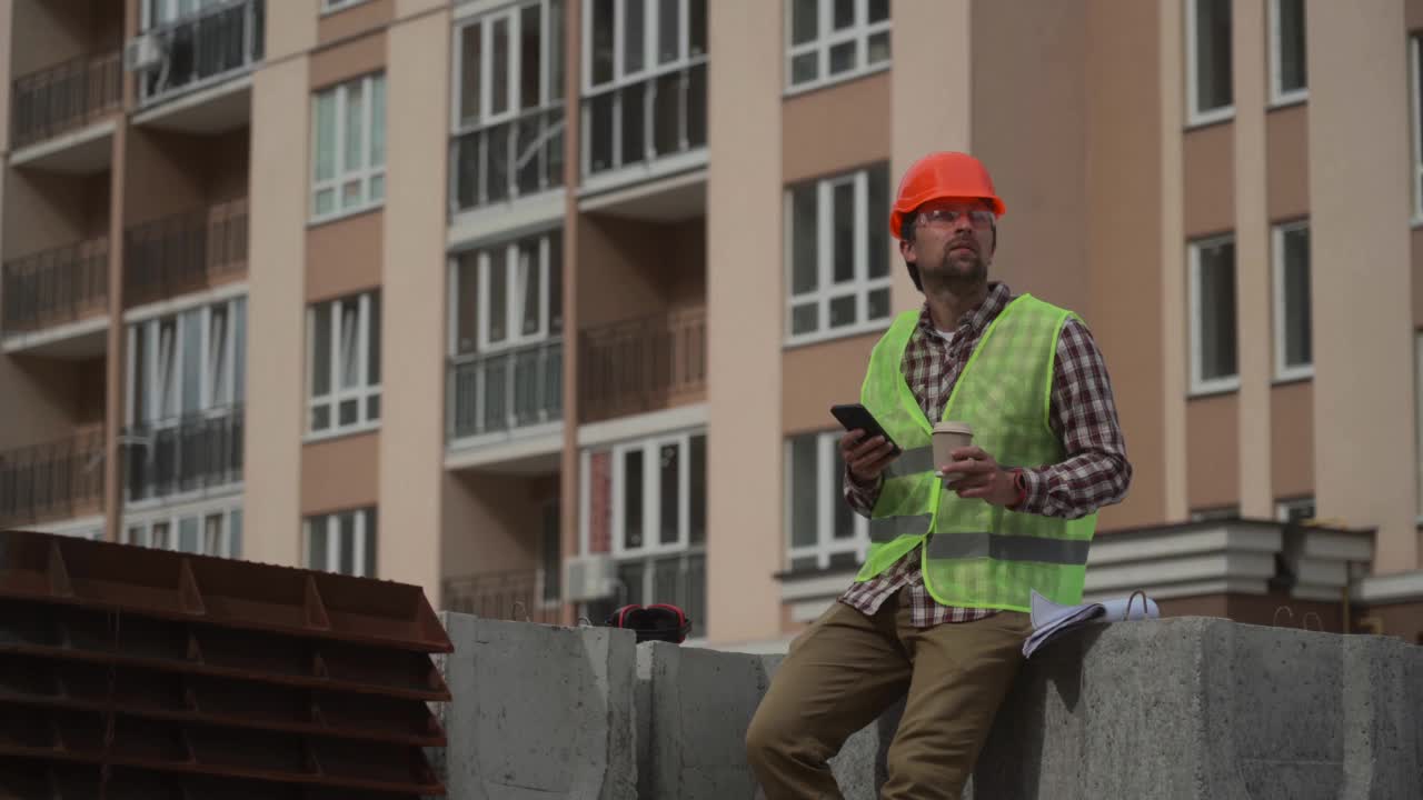 工头建筑工男年轻白人在工地休息休息期间喝着咖啡，看着手机，戴着安全帽，护目镜，反光背心。主题建构与建筑视频素材