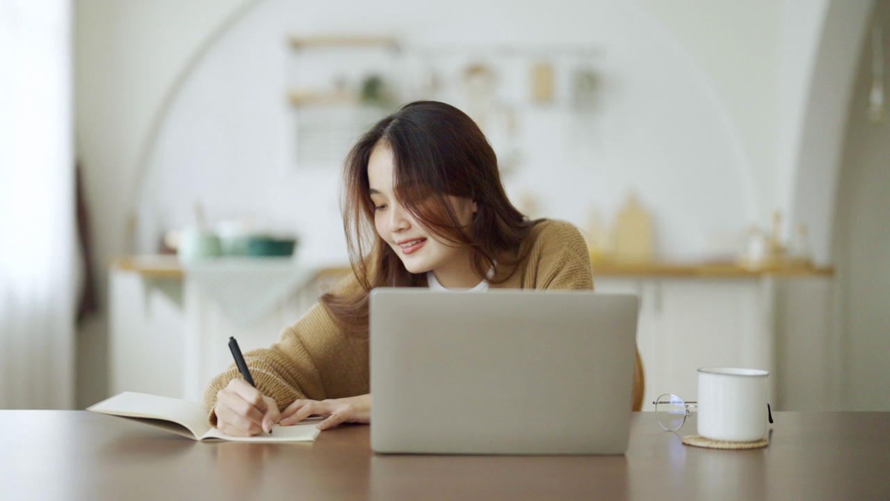 年轻的亚洲女人坐在桌子上，在笔记本上做笔记。在桌子上用笔记本电脑、文件和咖啡工作。学生学习在网上视频下载