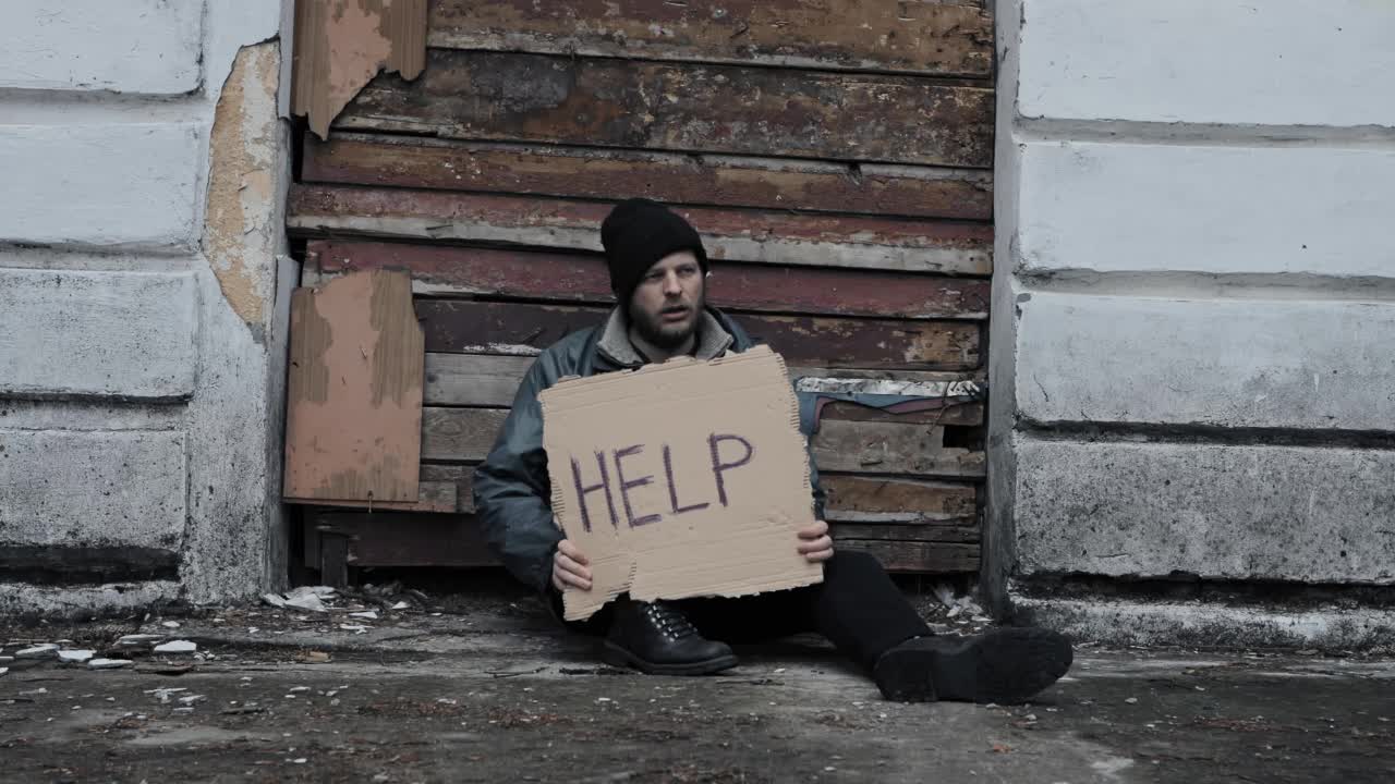 无家可归的人坐着，手里拿着一块写着“救命”的纸板。正在找工作的难民。穿着脏衣服，戴着帽子的流浪汉，生活在贫困线以下。大胡子逃亡的乞丐。移民。醉酒视频素材