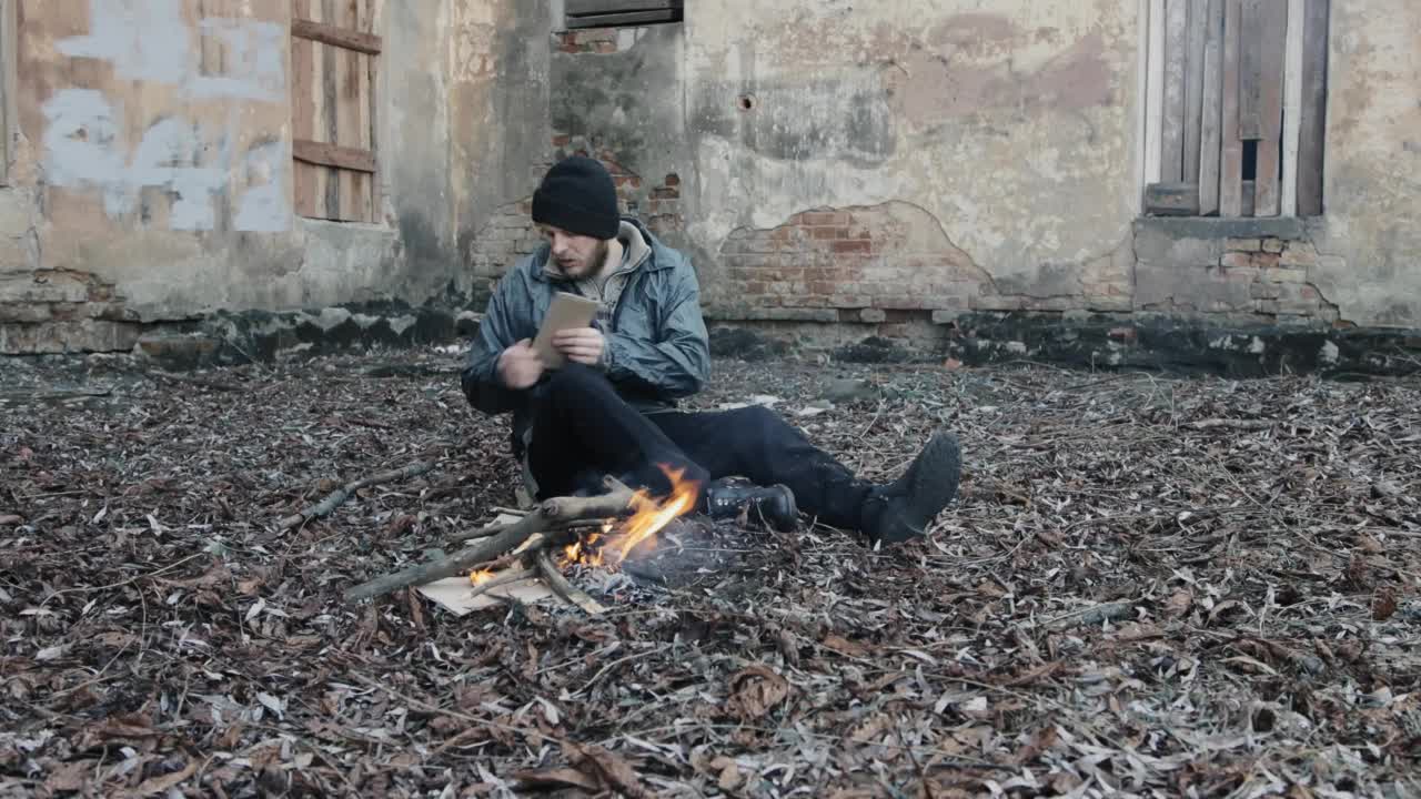 一个无家可归的人坐在地上，把几块硬纸板推入火中取暖。流浪汉穿着脏衣服，戴着帽子，生活在贫困线以下。非法移民。男性的难民。逃亡的乞丐视频素材