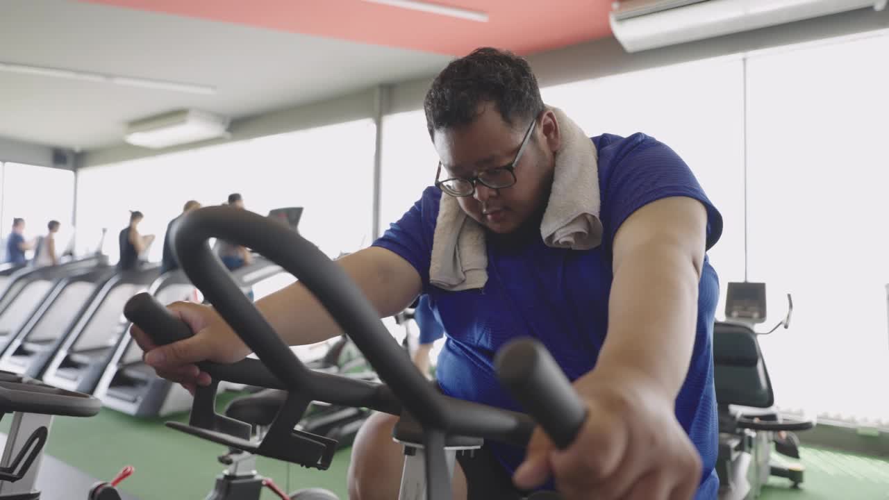 在健身房的健身课上，一名超重男子坚持在固定自行车上骑车。视频下载