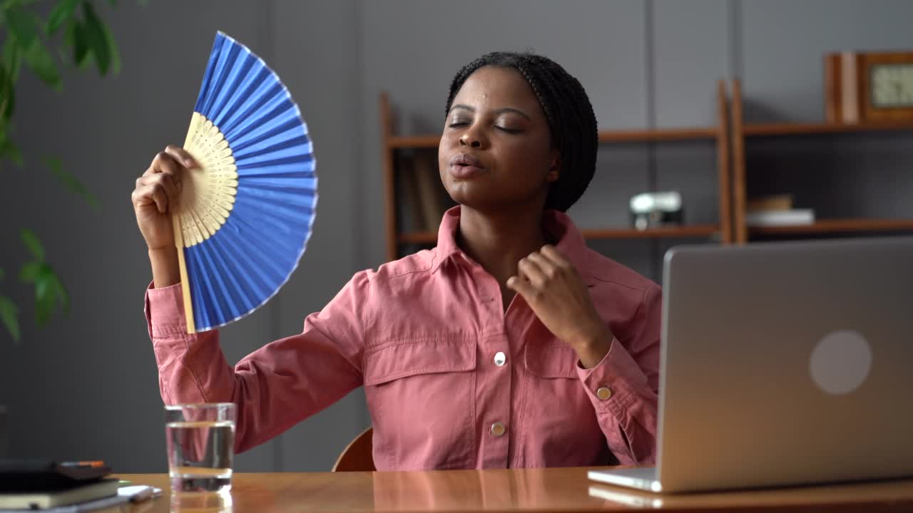 汗流浃背的非洲妇女工人坐在炎热的办公室笔记本电脑，试图避免过热使用纸扇视频素材