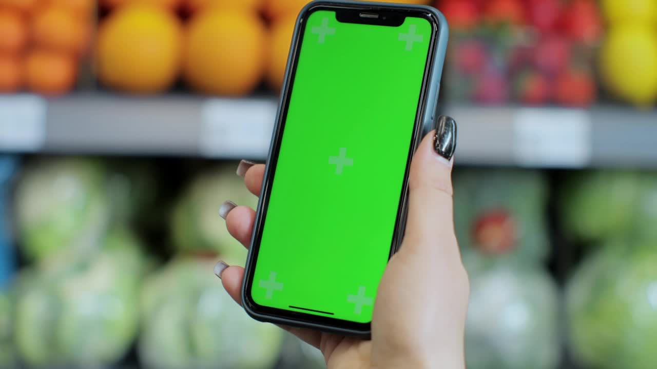 特写女人手握智能手机与绿色屏幕模糊背景水果和蔬菜在超市。视频素材