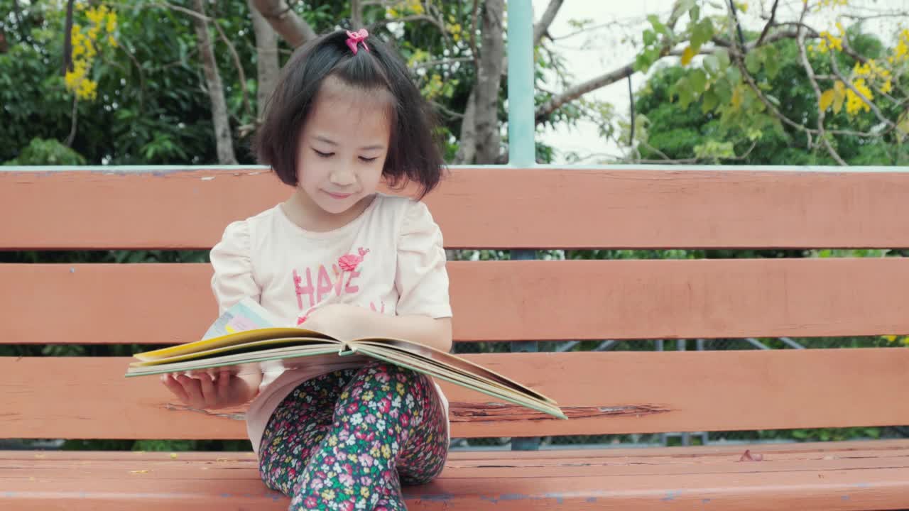可爱的亚洲女孩在学校户外独自看书，展示了自学和在夏天的闲暇时间学习的概念。它是孩子们的放松和享受。视频素材