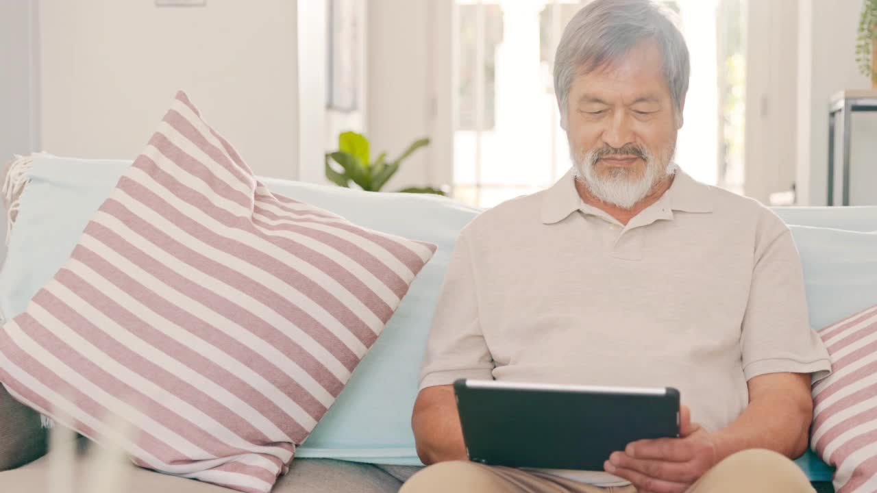 年长的亚洲人独自坐在家里的沙发上，使用数字平板电脑。一位亚洲老人正在客厅里用数码平板电脑上网视频素材