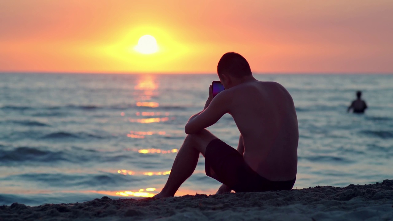 年轻人坐在热带沙滩上，用智能手机拍美丽的海滩日落视频素材