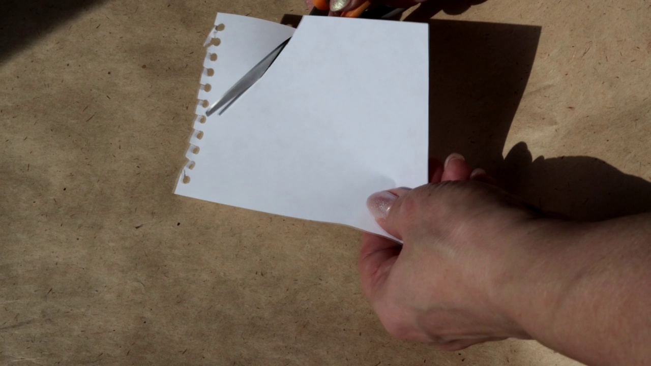 在一段4k视频中，一名女子用剪刀在棕色牛皮纸的背景上用手剪开一张空白的白纸视频下载