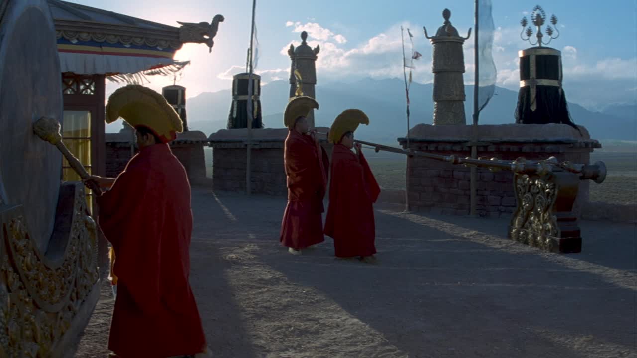 僧袍戴帽子的僧人在寺庙外演奏音乐的中等角度。右边的两个和尚吹长角，左边的和尚用大鼓槌敲大鼓。看看上面有大型宗教符号的石墙。看到山视频下载