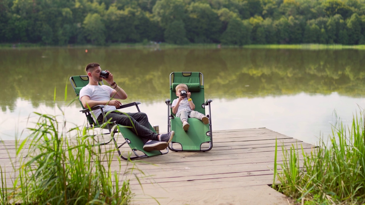 快乐的父亲和小儿子坐在湖边木制码头舒适的椅子上休息。爸爸和小男孩一起在户外度过休闲时光视频下载
