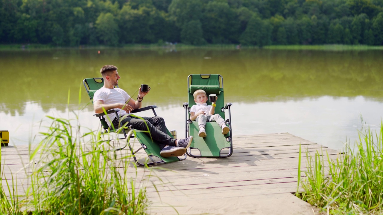 快乐的父亲和小儿子坐在湖边木制码头舒适的椅子上休息。爸爸和小男孩一起在户外度过闲暇时光视频下载