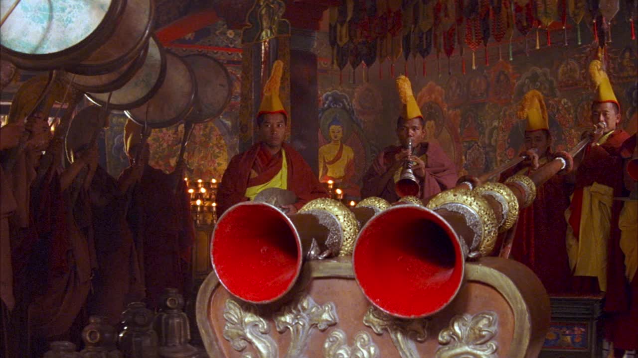 中等角度的僧侣穿着红色和金色的斗篷，戴着黄色的头饰。看和尚在吹小号、铙钹和鼓。看窗帘的边缘从头顶垂下来。背景中有蜡烛和壁画。可能是为了宗教仪式。视频下载