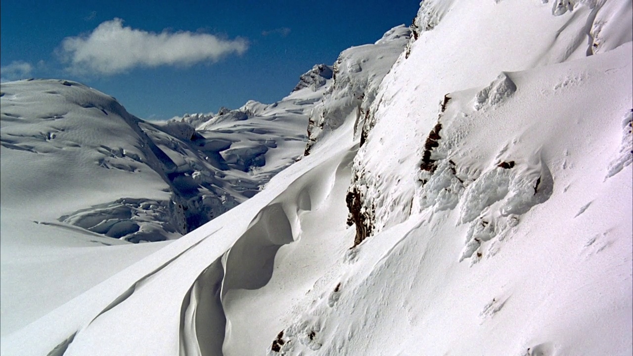 积雪覆盖的山坡。看到白雪覆盖的山坡或冰川在背景。在蓝色的天空中，左侧是单瓣积云。看顶部的阴影。可能是直升机叶片。视频素材