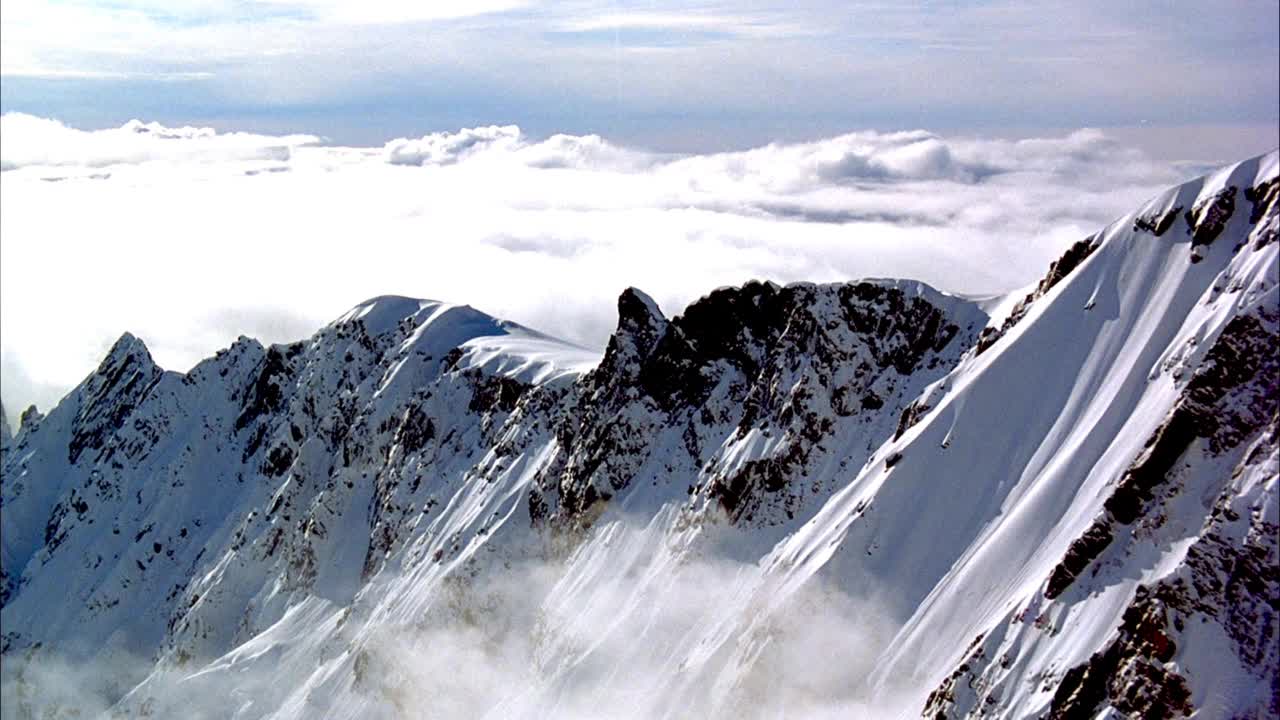 空中的雪山山峰从白色积云的海洋中窥见。在积雪覆盖的斜坡或冰川上滑行。向下滑行到积雪覆盖的山坡和布满云雾的冰川山谷。看到偶尔的镜头闪光从苏视频素材