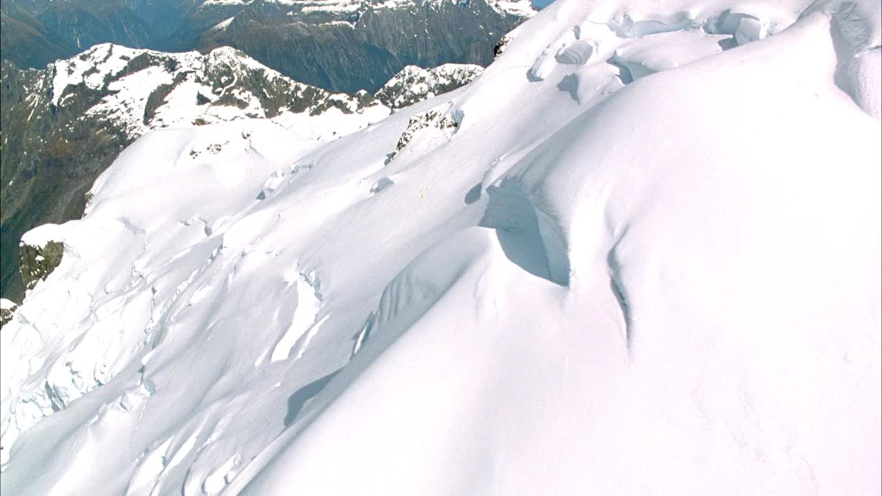 在白雪覆盖的山峰上架空。在山坡上看到可能的冰川形成。看到雪地上直升机的影子。看到云覆盖天空和偶尔的镜头耀斑从太阳。视频素材