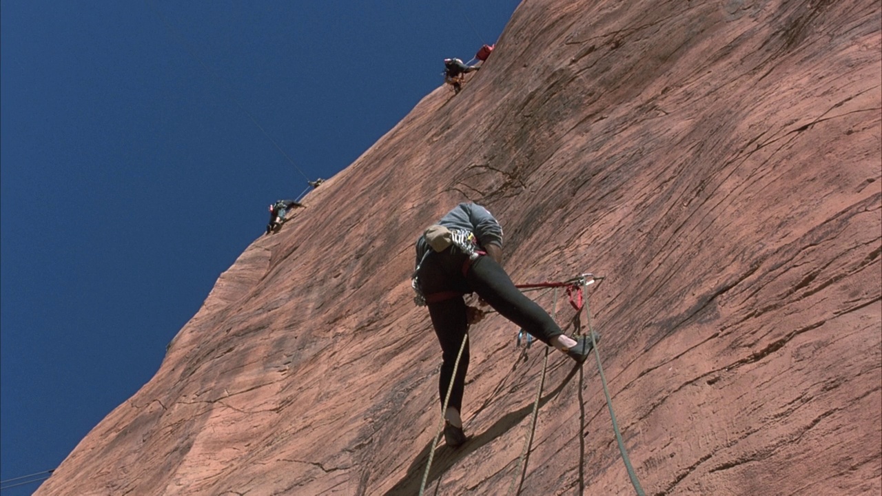 三名登山者用绳索支撑着生锈的彩色岩壁。看攀登者将绳子折叠起来并固定在墙上。在背景中看到清澈的蓝天。视频下载