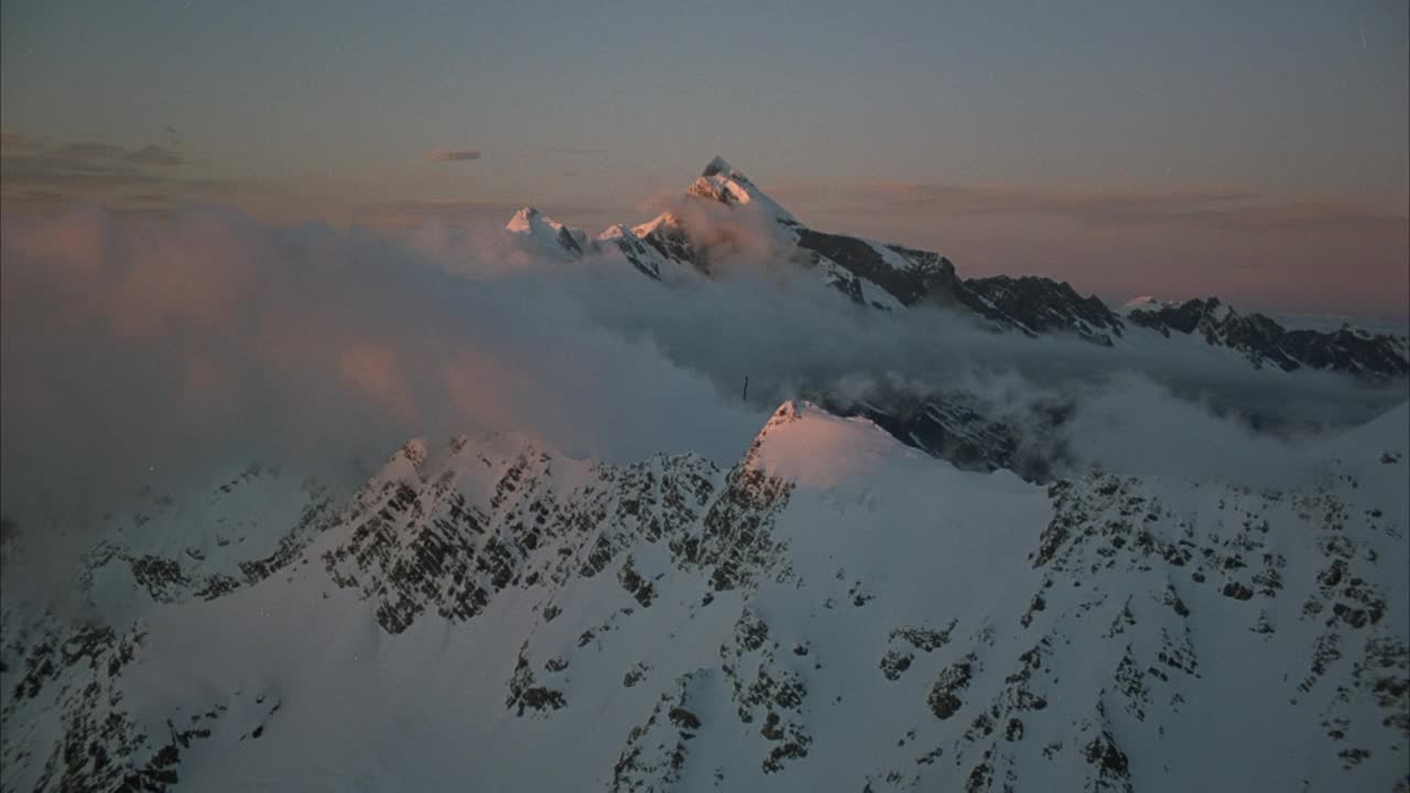 白雪覆盖的山顶在雪域之间的空中。可能是K2。看到最高的山峰下的云海，在前景中向上移动。在山峰周围和背景云上看到粉红色的色调。视频素材