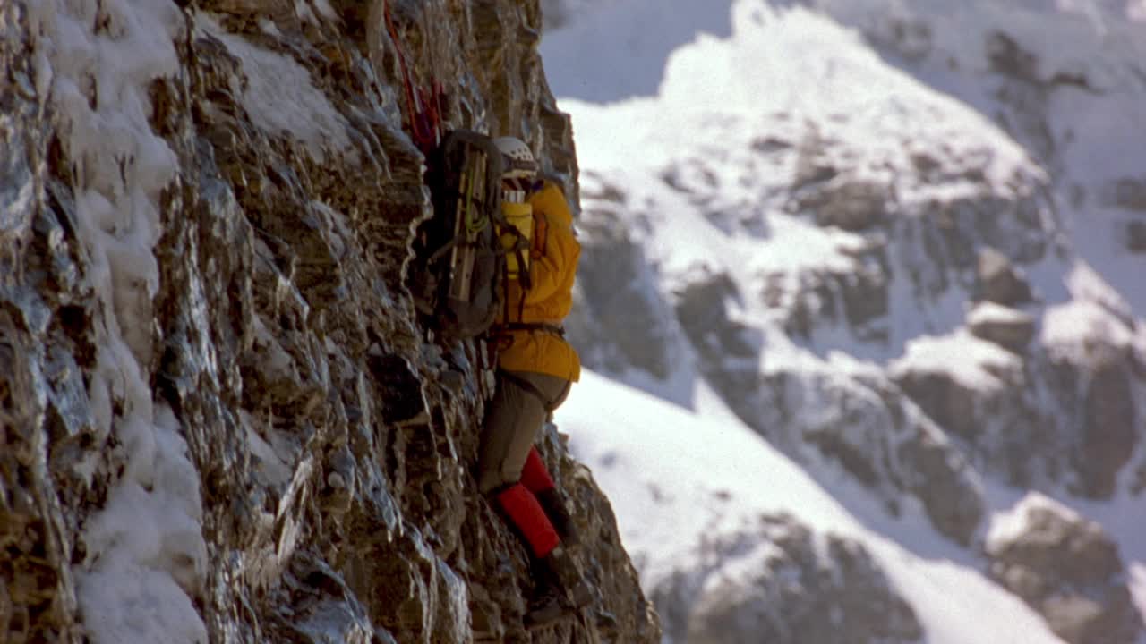中等角度的登山运动员紧抓着山坡、悬崖或布满积雪的岩壁。攀爬到第二个登山者，在把绳子固定到墙上时向下看了一眼。在背景中看到清澈的蓝天。视频素材