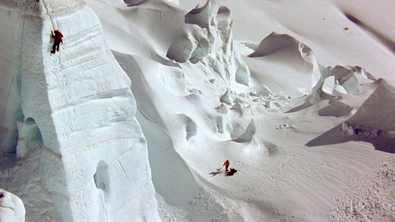 登山者在陡峭的白色冰或雪悬崖或墙壁上的空中拍摄。当登山者上山时，用斧子和雪鞋拉回来。看到观察员或第二个登山者站在悬崖底部的雪中观看，并抓住系在fi的绳子视频素材