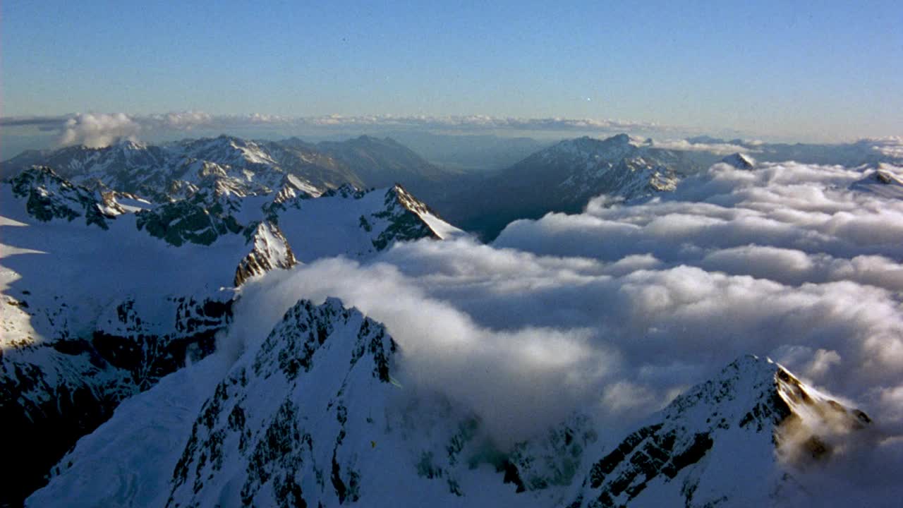 白雪皑皑的山脉。看到山顶与白色积云毯子在底部和周围的山脉。看波峰从云天的一边飘到另一边。看到有光晕和透镜耀斑的太阳。见直升机尾部。视频素材