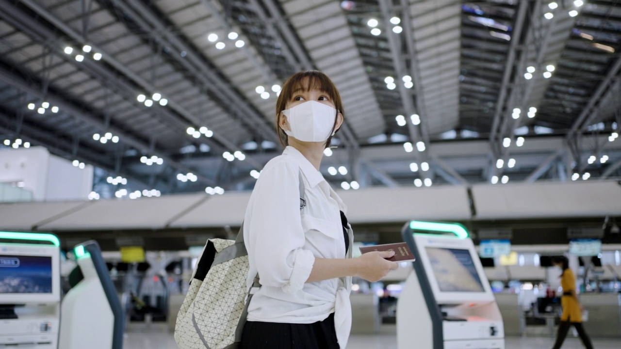 一名亚洲年轻女子戴着防护口罩，拿着行李在机场候机楼走着视频素材