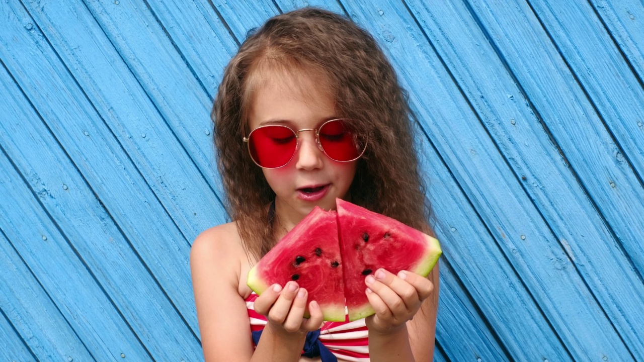 一个卷发的黑发小女孩手里拿着两块红色的西瓜。一个孩子喜欢在热天咬下一片多汁的甜西瓜视频下载