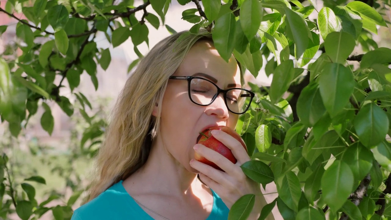 一个年轻女人咬着吃着一个成熟的红苹果，看着花园里的摄像机，慢动作。健康饮食和美容。饮食。视频素材