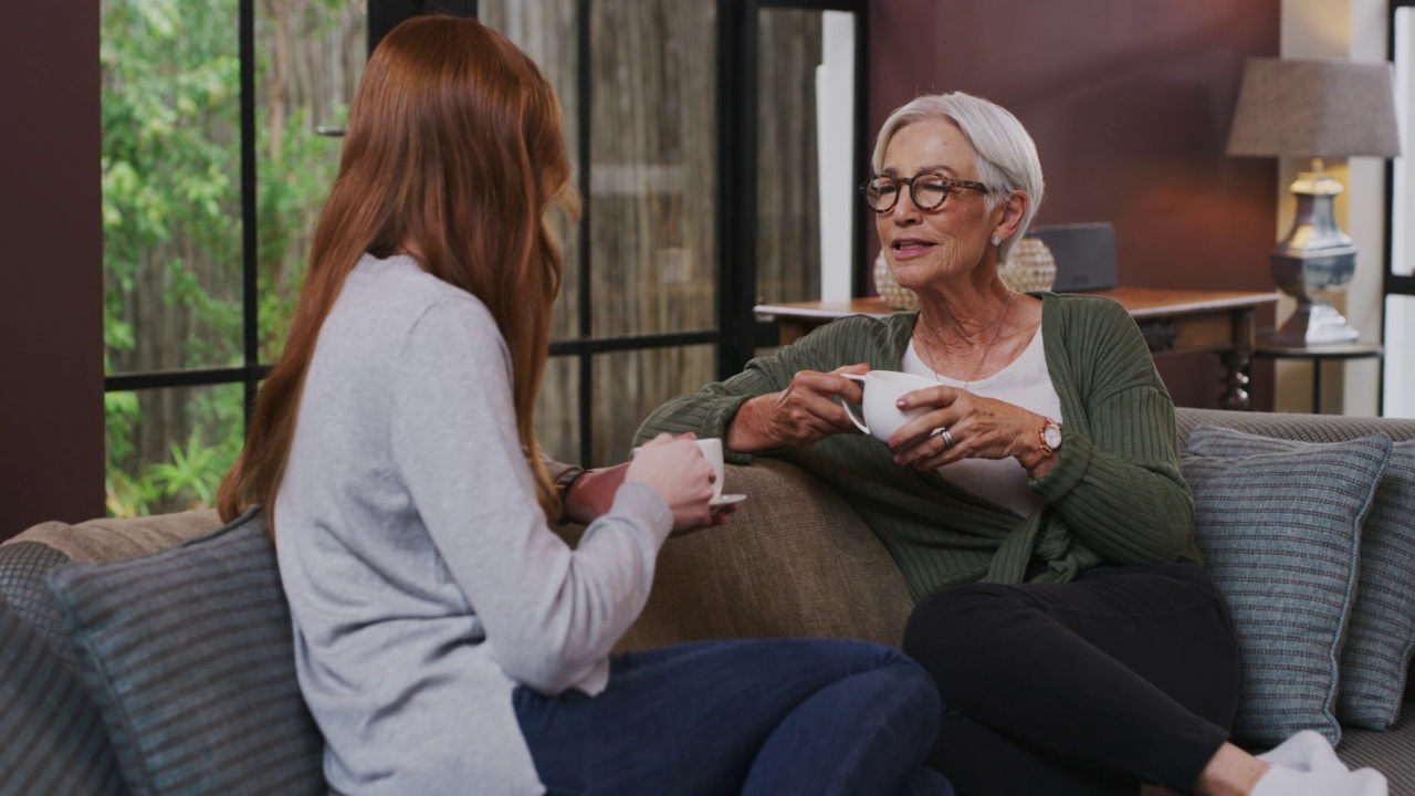 一个年轻的女人在家里喝着咖啡，和她年迈的母亲聊天。老太太在家里和女儿聊天，喝茶视频素材